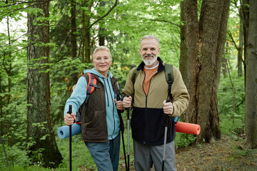 Senior Caucasian woman and senior Caucasian man standing in forest holding trekking poles and yoga...