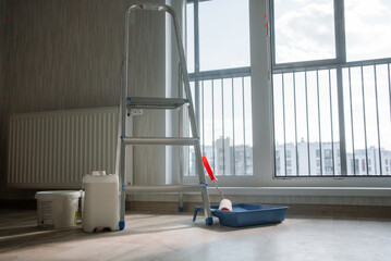 Metal stepladder positioned near a paint roller, tray, and buckets, indicating an upcoming...