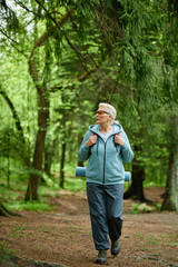 Senior Caucasian woman walking through forest carrying backpack and yoga mat, wearing sunglasses,...