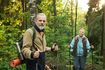 Caucasian senior man and Caucasian senior woman hiking with trekking poles in forest, carrying...