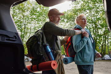 Senior Caucasian man helping senior Caucasian woman adjust backpack while preparing for outdoor...