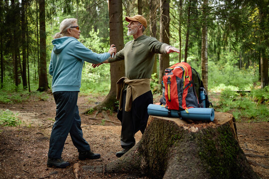 Senior Caucasian man standing in forest with arms outstretched while senior Caucasian woman holding his hand, both smiling and interacting near large backpack on tree stump
