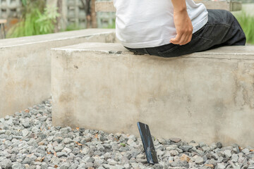 A man sits on a cement bench. The bench is next to a wall. The man's cell phone falls.