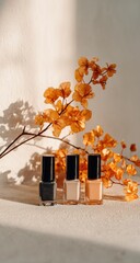 Three nail polishes in muted tones sit beside dried orange flowers, bathed in sunlight against a textured white backdrop