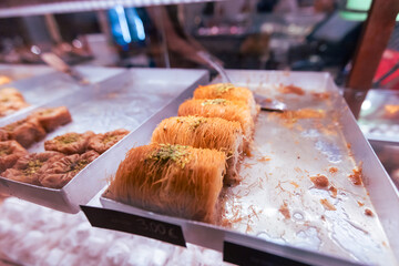 Golden kunafa shredded phyllo pastries topped with crushed pistachios and soaked in honey syrup, beautifully arranged in professional metal display trays at authentic Middle Eastern sweet shop.
