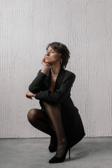 woman crouches elegantly in a dark suit with sheer stockings, resting her chin on her hand, showcasing a confident and contemplative expression in a modern studio