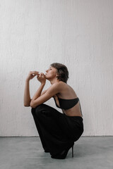 model showcases a stylish black outfit while crouching gracefully against a textured wall. minimalist setting emphasizes her pose and fashion aesthetic during the shoot