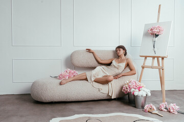 woman lounges elegantly on a unique sofa, surrounded by pink flowers and an easel displaying floral art in a bright and stylish interior space