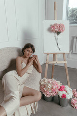 woman in a sleek, light-colored dress sits gracefully on a cream chair beside painted floral art. Soft pastel flowers in buckets add color to the bright, airy studio
