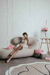 graceful woman sits relaxed on a stylish sofa in a well-lit studio. She is surrounded by vibrant flowers and an easel, suggesting a creative atmosphere
