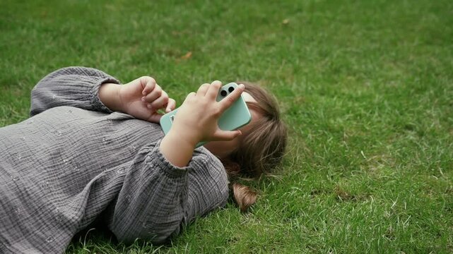 Child girl lying on the grass in a garden, using a smartphone.
