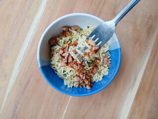 a bowl of instant noodles topped with chopped chicken, spring onions and fried shallots on a wooden...
