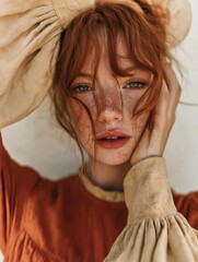 Close-up portrait of a young woman with red hair and freckles on her face. she is wearing a beige hat with a wide brim and a long-sleeved top with a ruffled neckline.