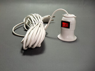 An electrical fixture with a coiled white cable and plug rests on a neutral gray surface, featuring a red on/off switch, ready for providing power and illumination.