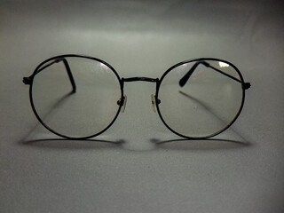 Round eyeglasses resting on a textured gray surface, displaying a classic and intellectual aesthetic, the clear lenses reflecting subtle light in studio lighting.