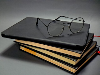 A stack of black journals and notebooks with round glasses resting on the top journal, rendered with natural light and dark shadows emphasizing the textural details.