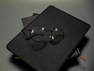 Elegant round spectacles are artfully placed on a dark sketchbook sitting atop more books against a muted grey backdrop, creating a sophisticated and scholarly visual statement.