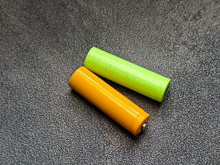 A high-angle shot reveals two cylindrical batteries, one golden yellow and the other light green, placed diagonally on a textured gray surface, casting subtle reflections of light.