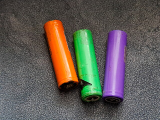 A trio of colorful cylindrical cells lies side-by-side, showcasing a vibrant orange, a lively green with visible damage, and a pristine purple, all resting on a textured surface.