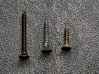 Three screws of varying sizes and colors are displayed on a textured gray surface, showcasing the tools used in construction and repair projects in industrial contexts.