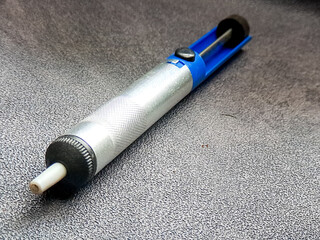 Silver desoldering pump tool showing functional design and utility, with blue accents against a textured surface, showcasing precision and practical craftsmanship.