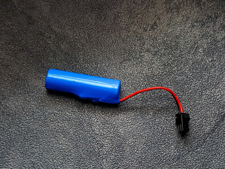 Vivid cylindrical battery with a bright blue casing, connected by a sleek red wire to a small black connector, rests on a textured dark surface showcasing functional design.