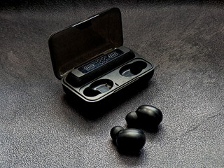 Black wireless earbuds rest near their partially open charging case, displaying battery levels and sleek design, set against a textured dark gray backdrop, conveying modernity.