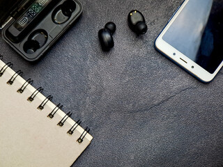 Top-down composition featuring a spiral-bound notebook, cordless earbuds, a charging case and smartphone on a textured dark surface conveys technology and analog elements together.