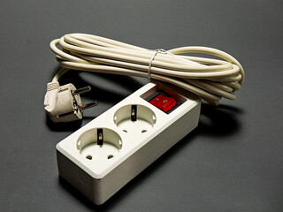 A white power strip with two outlets and coiled cord sits on a gray surface, the red switch is on indicating the flow of electricity, a common appliance for power distribution.
