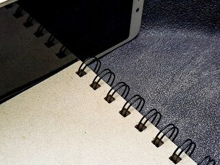 A spiral notebook with black rings sits partially under a device, placed atop a textured surface, highlighting the simple tools of organization and digital integration.