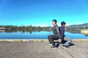 Fototapeta premium Two Men Perform A Partner Squat Outdoors, Holding Hands And Leaning Back To Back By A Peaceful Lake