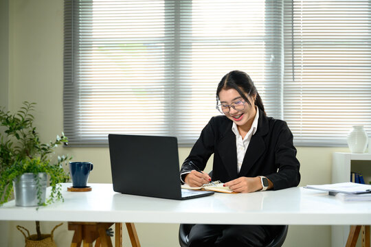 Smiling Asian businesswoman using a laptop and writing in a notebook - Powered by Adobe