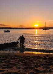  serene sunset over a tranquil beach, capturing a vibrant orange sun reflecting across calm waters. A silhouette of a fisherman and his dinghy stands in the foreground, with soft sand adding texture. 