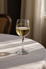 Glass of wine standing on round table near window with sunlight casting shadows across surface, background featuring blurred chair and light curtains