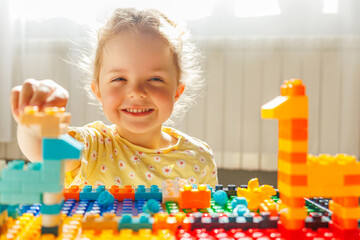 
A beaming toddler girl with a messy bun smiles brightly at the camera while playing with colorful...