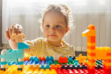 A happy toddler girl with a messy bun smiles while playing with colorful building blocks, focused...