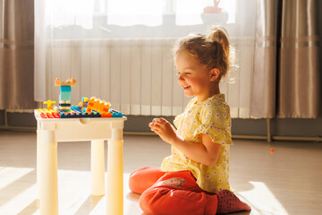 A cheerful toddler girl sits on the floor, smiling happily as she plays with colorful building...