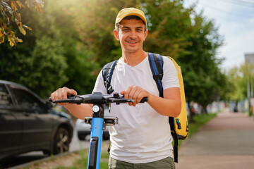 Smiling delivery courier with a yellow backpack rides an electric scooter on a sunny day. Fast and...