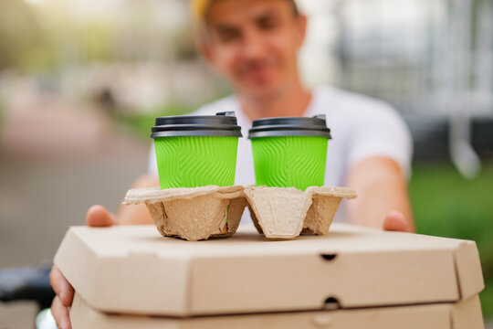 A smiling delivery man holds out a stack of pizza boxes and two green coffee cups in a cardboard carrier. The focus is on the food and drinks, with the man suggesting fast and reliable service.