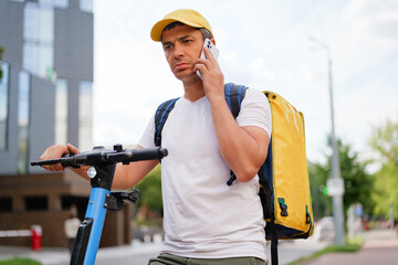 A delivery man with a backpack on a scooter talks on his phone with a serious expression, listening...