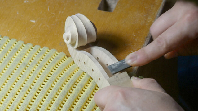 Skilled luthier carefully carving wooden violin neck with specialized chisel, highlighting intricate craftsmanship of traditional instrument making