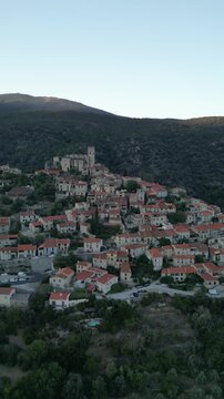Aerial view of sunlit Eus village, perched on a lush hillside with terracotta rooftops and a prominent church tower, Eus, Occitanie, France.