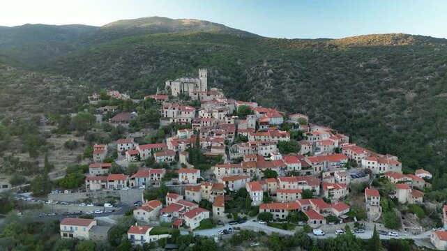 Aerial view of Eus village with its terracotta rooftops cascading down the hillside crowned by the church amidst lush green hills, Eus, Occitanie, France.