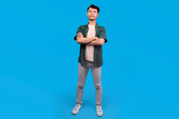 Portrait Of A Young Man With His Arms Crossed, Wearing A Green Shirt Against Blue Studio Background