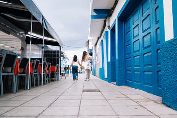 A woman and two children walk down a street in front of a blue building in Agaete Gran Canaria. The...