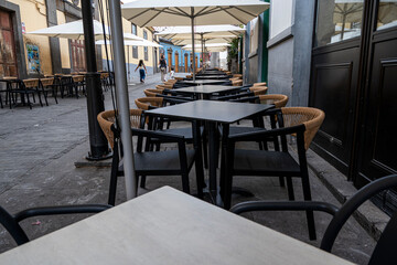 A row of tables and chairs are empty in a restaurant. The tables are covered with white tablecloths and the chairs are black. The scene is quiet and empty
