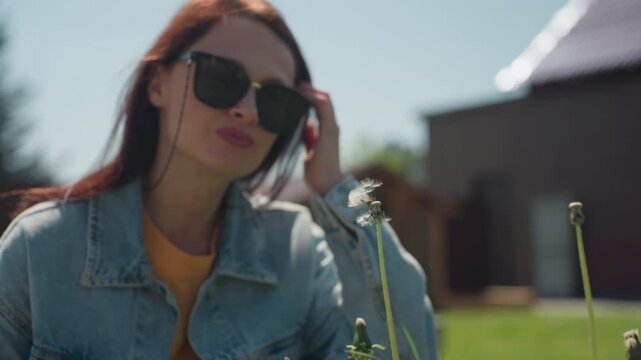 Close up of delicate dandelion with white cotton seeds ready to disperse, with woman bending in background preparing to blow on it, set in sunny green backyard with house and garden in soft blur