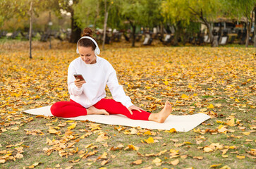 Outdoors woman lifestyle. Young female in headphones checking smartphone on yoga mat in fall park