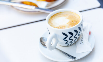 A cup of coffee with milk. On the saucer are two sugar packets and a spoon.