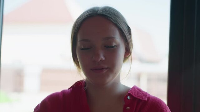 Lady in red shirt shyly covers mouth with hands, reacting with playful and expressive face while interacting with unseen people in background, bright indoor light adding warmth to candid moment